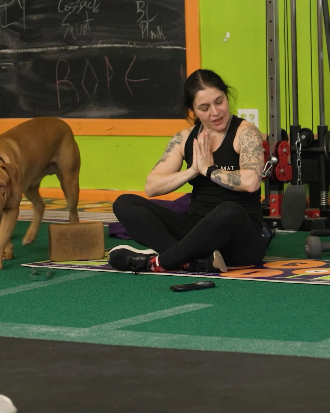 A member seated cross-legged on the turf with hands together, a dog behind her